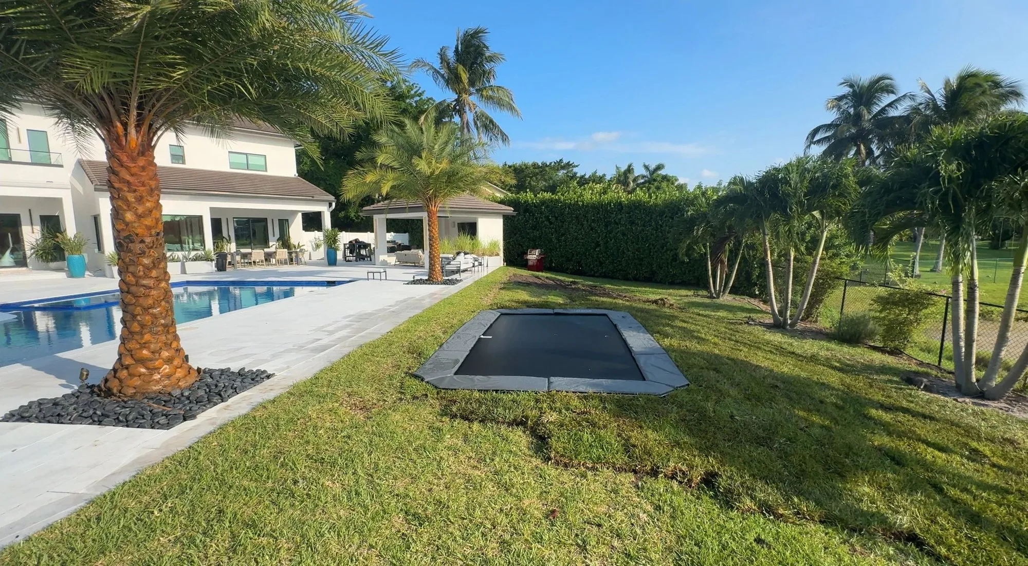 Rectangular in-ground trampoline set flush in a newly laid lawn beside a pool deck, palms and a white two-story house
