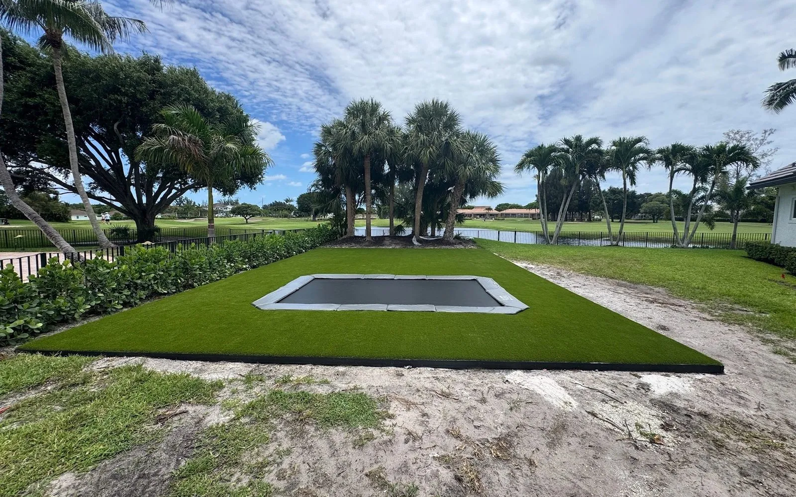 A rectangle in-ground trampoline sitting flush in a new artificial turf surround, with the ground around it still bare sand from the dig
