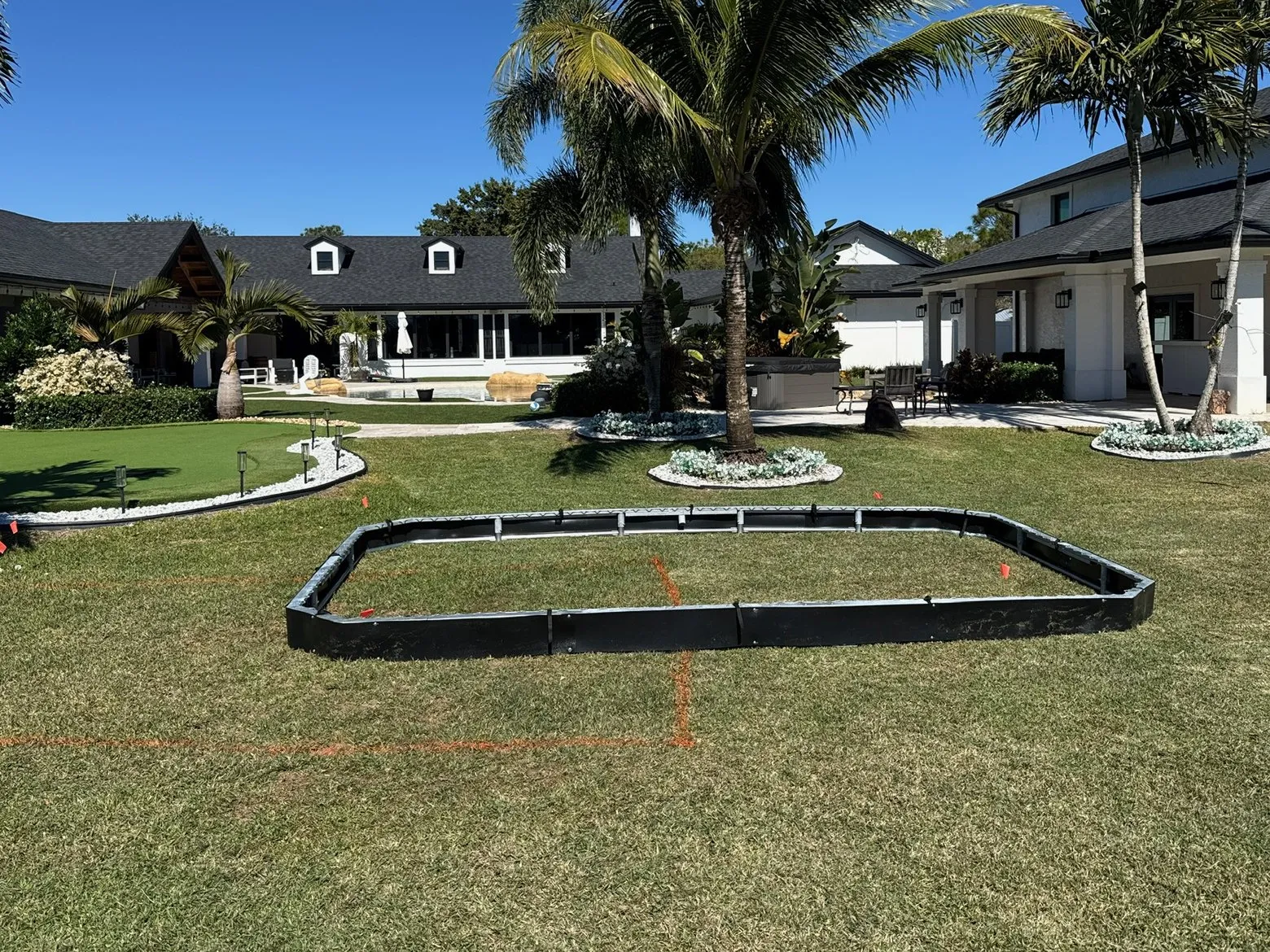 Before: a backyard lawn with the retaining wall laid out on the grass where the trampoline will go, palms and a white house behind