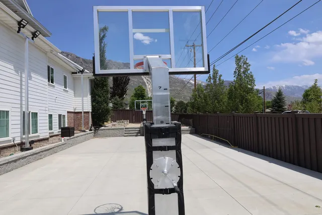 An installed 60 inch hoop seen from below and behind the baseline, the clear board against a bright sky with a white two storey house and mountains behind it.