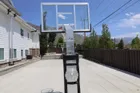 An installed 60 inch hoop seen from below and behind the baseline, the clear board against a bright sky with a white two storey house and mountains behind it.
