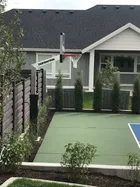 A 60 inch hoop standing at the end of a green home court beside a house, with the pole padded in black and a hedge of young trees along the fence line.