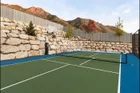 A 60 inch hoop at the far end of a green and blue home court, set against a stacked boulder retaining wall with a pickleball net across the court and mountains behind.