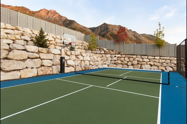 A 60 inch hoop at the far end of a green and blue home court, set against a stacked boulder retaining wall with a pickleball net across the court and mountains behind.