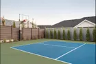 A 60 inch hoop in the corner of a fenced green and blue court beside a new house, its pole padded in black and a row of young cypress trees along the fence.