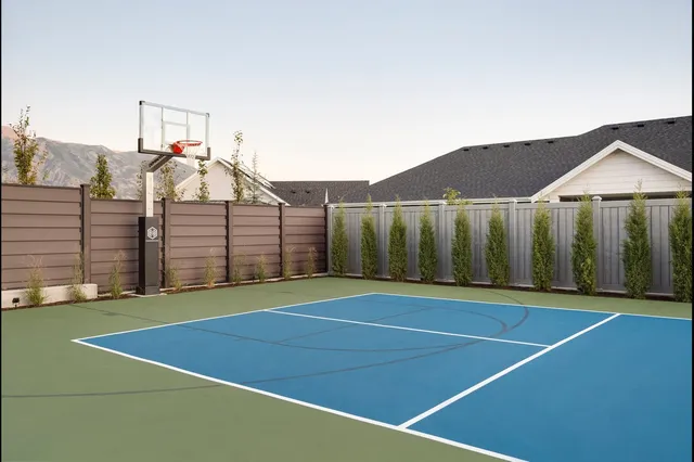 A 60 inch hoop in the corner of a fenced green and blue court beside a new house, its pole padded in black and a row of young cypress trees along the fence.