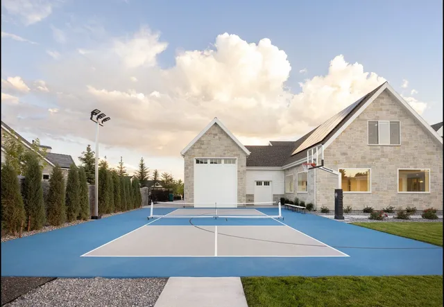 The 72 inch hoop installed at the end of a pale blue home court, seen from the far baseline with a stone house and light poles behind it, showing how far back the four foot overhang keeps the pole.