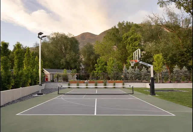 A 72 inch hoop at the side of a grey and green court with a pickleball net, a light pole in the far corner and a landscaped garden behind it.