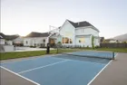 A 72 inch hoop at the end of a blue pickleball court in front of a large white house, the net strung across the court in the foreground.