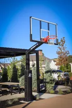 A black 72 inch hoop at the end of a home court beside a pergola and outdoor dining table, the dark frame sitting against the trees rather than standing out from them.