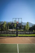 A black 72 inch hoop seen straight on from the far baseline of a tan and green home court, with a large house and mountains behind it.
