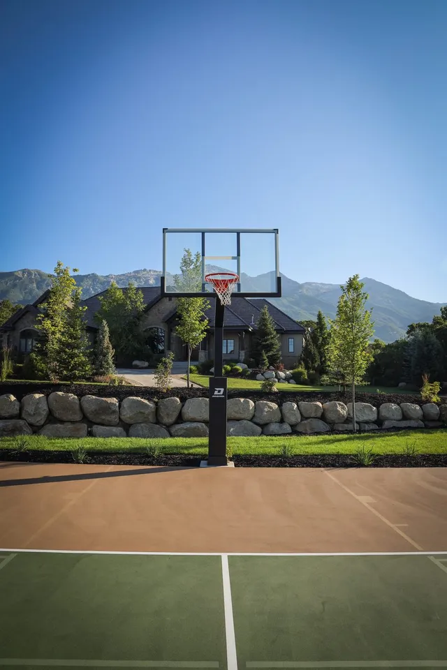 A black 72 inch hoop seen straight on from the far baseline of a tan and green home court, with a large house and mountains behind it.