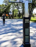 A fixed height hoop installed on a public park court, seen close along the padded post with a second hoop, a playground and trees further down the court.