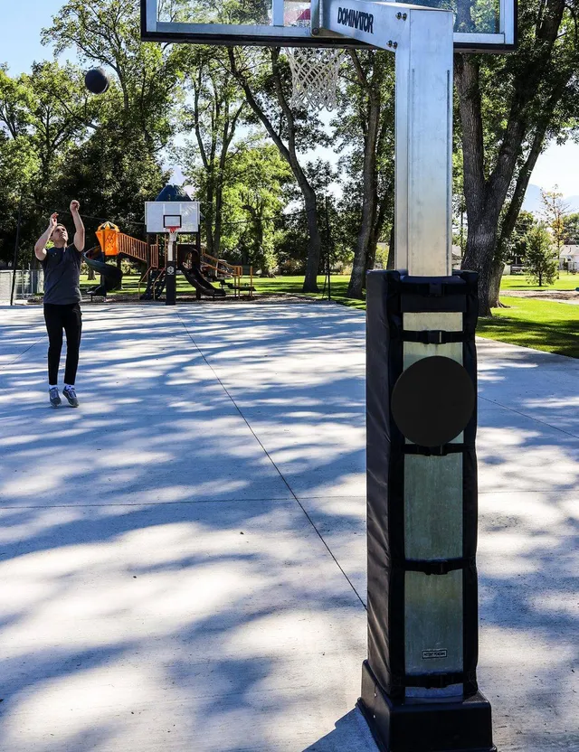A fixed height hoop installed on a public park court, seen close along the padded post with a second hoop, a playground and trees further down the court.