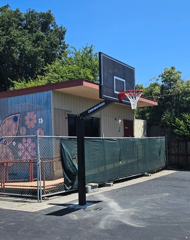 A fixed height hoop on a school blacktop against a painted mural wall, the black post set straight into the asphalt with no anchor plate showing.