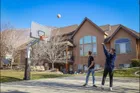 Two people shooting at a 72 Pro on a concrete court in front of a large stucco house, the ball in the air above the board.