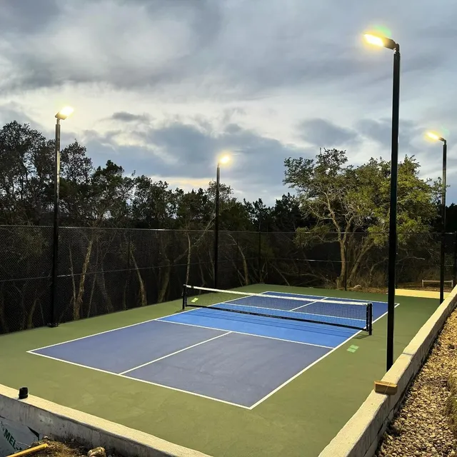 A blue and grey home court at dusk with its pole lights on, four poles around the court and the pickleball net up across the middle.