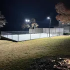 A lit court at night seen from the grass outside the fence, the pole lights throwing an even sheet of light across the whole surface.