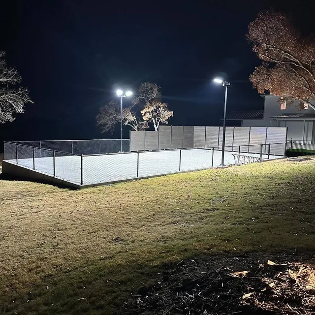 A lit court at night seen from the grass outside the fence, the pole lights throwing an even sheet of light across the whole surface.