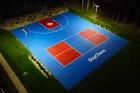 An aerial view at night of a blue and red court lit by pole lights at its corners, the light reaching the far corners of the surface.