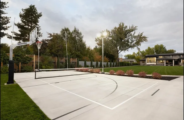 A two head light pole lit at the side of a concrete court on an overcast evening, with a hoop and a pickleball net on the court and a lawn beyond.