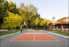 A three head light pole in the corner of an orange and grey pickleball court beside a modern house, with a basketball hoop at the far end.