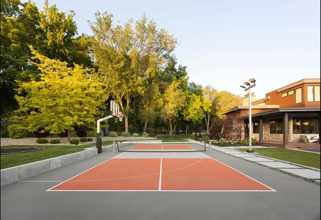A three head light pole in the corner of an orange and grey pickleball court beside a modern house, with a basketball hoop at the far end.