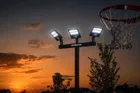 A black three head light pole lit against an orange sunset sky, the Dominator wordmark on the crossbar and a basketball net in the corner of the frame.