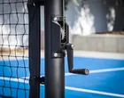 Close on the black Pro post, showing the winder mechanism and its crank handle with the net tape running off to the left and blue court beyond.