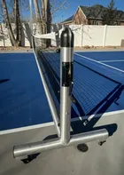 The Pro net set up on a blue outdoor court, photographed close along the frame so the round post, the winder and the locking caster underneath it are all in one frame.