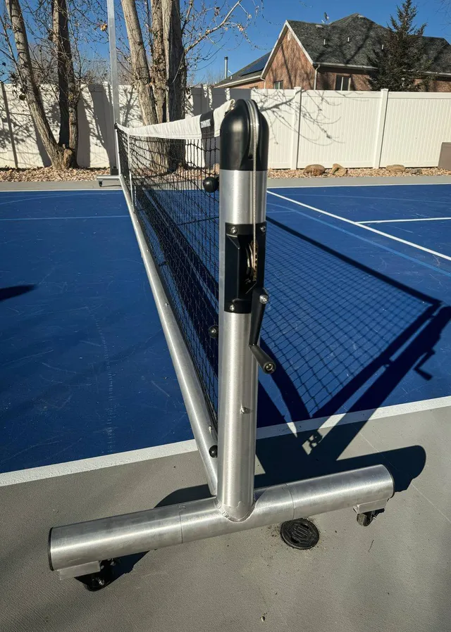 The Pro net set up on a blue outdoor court, photographed close along the frame so the round post, the winder and the locking caster underneath it are all in one frame.