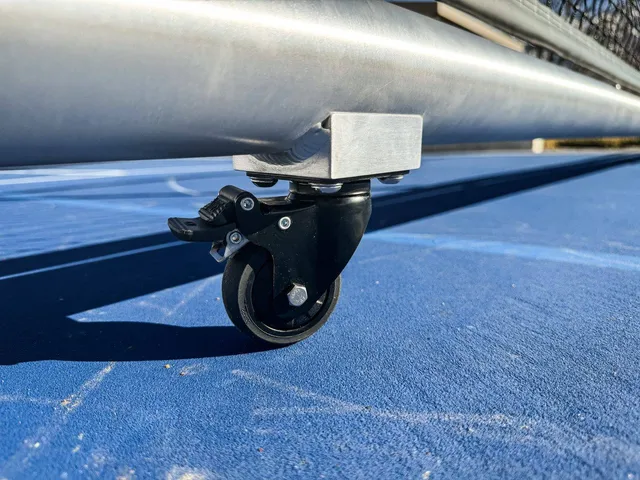 Close on one locking caster of the Pro net, the brushed round frame above it and the painted court line running underneath.