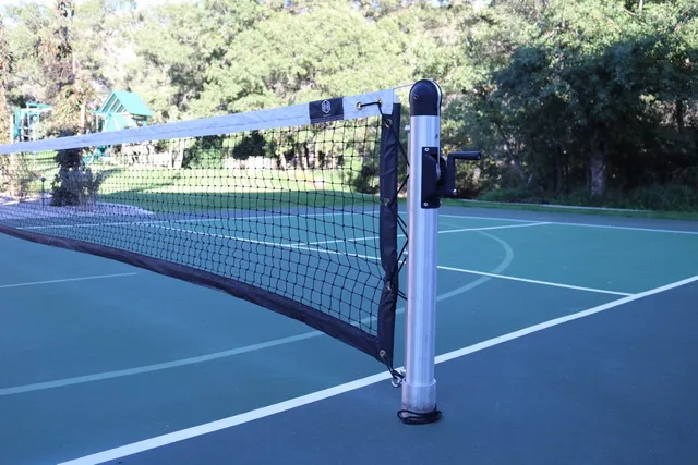 A round in-ground post installed on a blue and green court, the net running away from it and the collar of the ground sleeve visible where the post meets the surface.