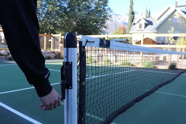 A hand turning the crank handle on an installed round post to tension the net across the court.