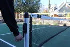 A hand turning the crank handle on an installed round post to tension the net across the court.