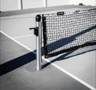 A round in-ground post set in its ground sleeve on a grey court, the crank handle on its side and the net tensioned away from it.