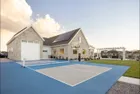 A Standard portable net set up across a blue and grey court in front of a stone house, with a basketball hoop by the garage.