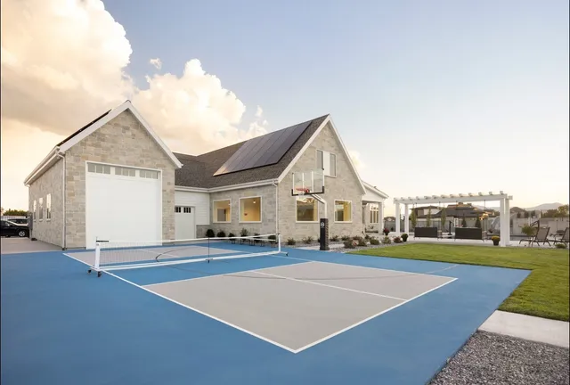 A Standard portable net set up across a blue and grey court in front of a stone house, with a basketball hoop by the garage.