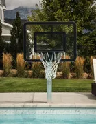 A poolside hoop installed at the edge of a swimming pool, the board overhanging the water on a short brushed aluminum pole set into the deck, with a lawn and mountains behind.