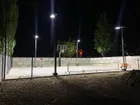 A court at night lit by solar poles, three lit heads standing above a fence line with the surface bright underneath them.
