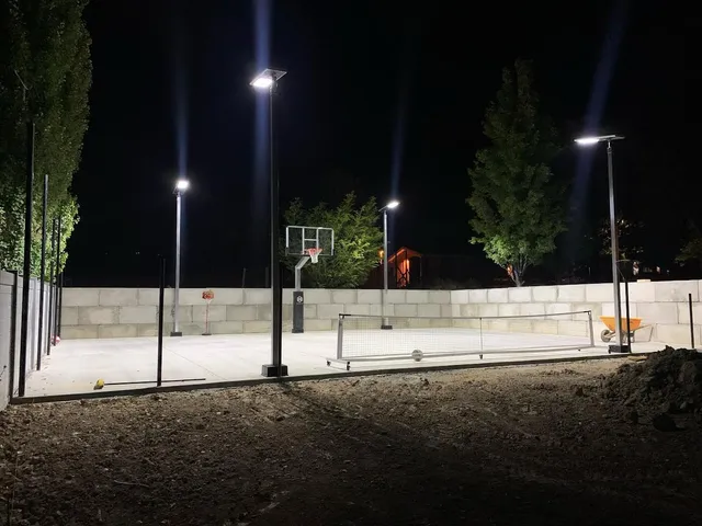A court at night lit by solar poles, three lit heads standing above a fence line with the surface bright underneath them.