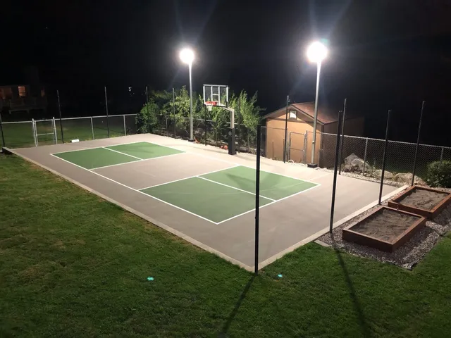 A green and tan court at night with two solar poles lit at one side, the light reaching across the whole playing surface.