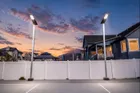 Two single head solar light poles lit at dusk along the white fence of a home court, with houses and a pink and blue sky behind.