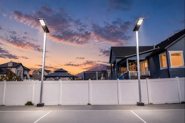 Two single head solar light poles lit at dusk along the white fence of a home court, with houses and a pink and blue sky behind.