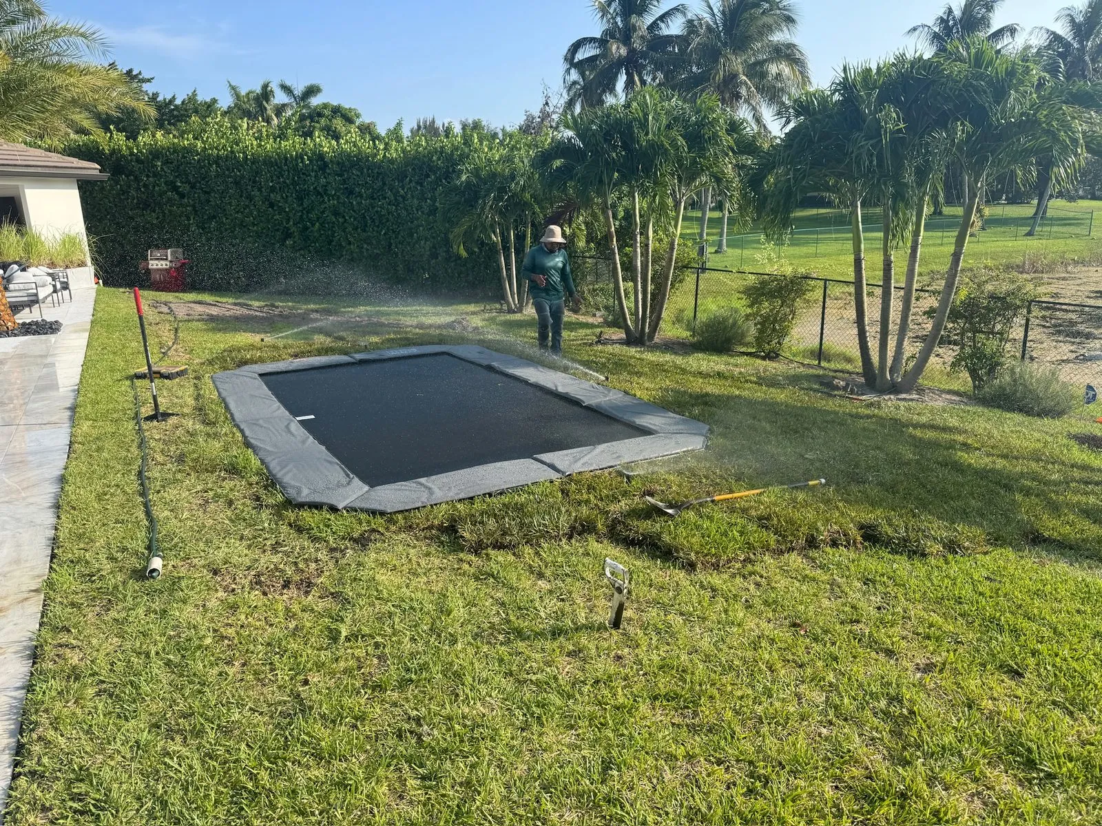 A rectangle in-ground trampoline set flush in a lawn, sprinklers running beside it, with a hedge and palms behind and a crew member at the far end
