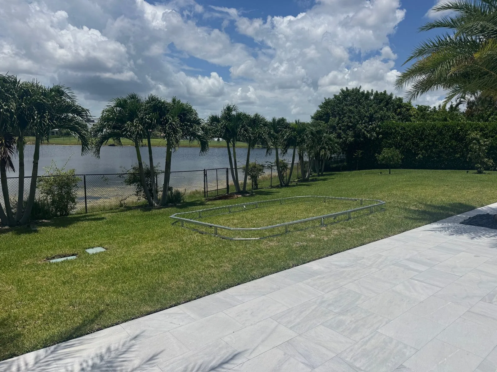 A rectangular metal frame laid out on a green lawn beside a white paver walk, with a chain-link fence, palms and a lake behind