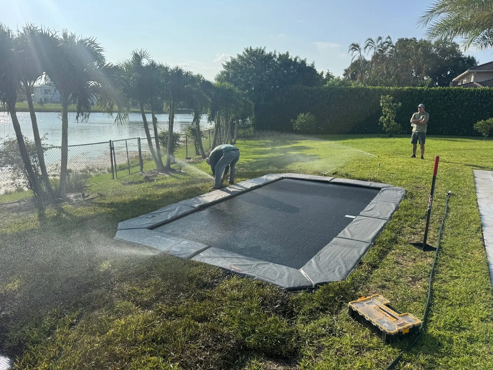 A rectangle in-ground trampoline in a freshly patched lawn with sprinklers running across it, a crew member kneeling at the pad and another standing by the hedge