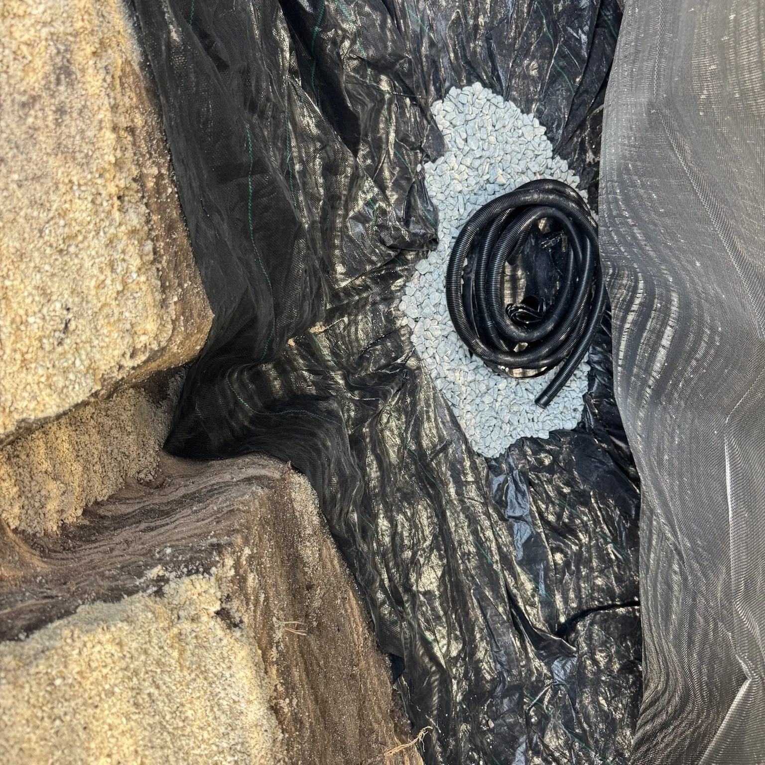 Inside a lined pit: a coiled black hose resting in a patch of pale gravel on black fabric, between the sandy wall of the dig and a sheet of liner