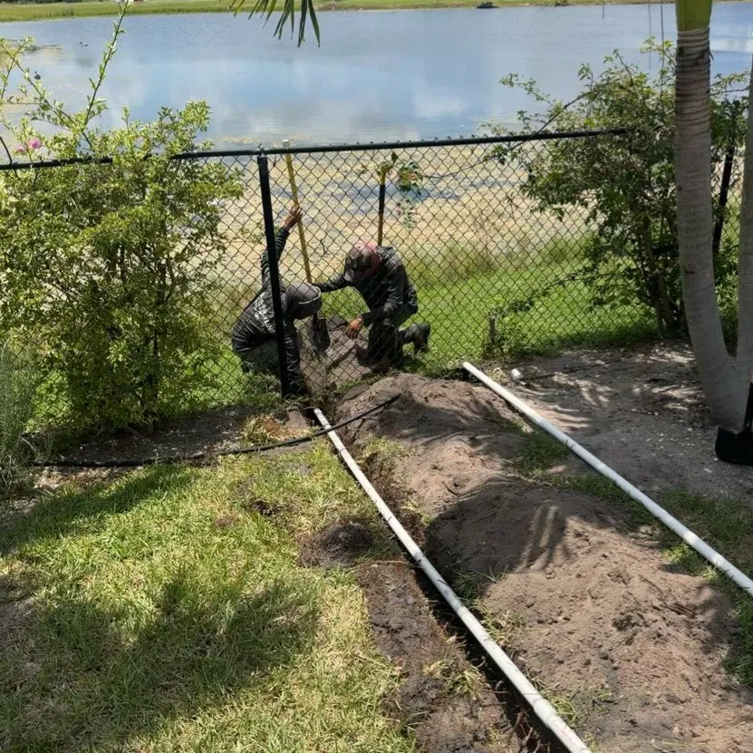 Two crew members kneeling at a chain-link fence beside a lake, with a fresh trench and lengths of white pipe across the lawn in front of them