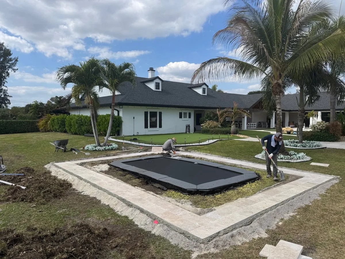 A rectangle in-ground trampoline set in its pit while two workers lay a border of pale pavers around it, with palms and a white house behind